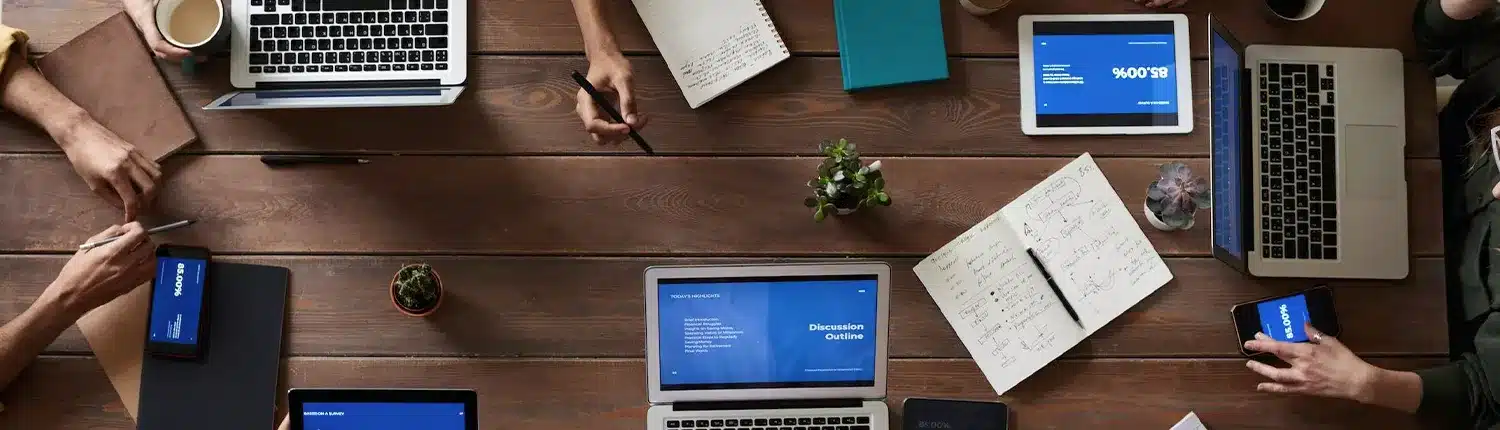 bovenaanzicht van tafel met laptops en mensen die daar aan werken, blaadjes agenda's, telefoons pennen in de hand en andere devices op tafel en in de hand.
